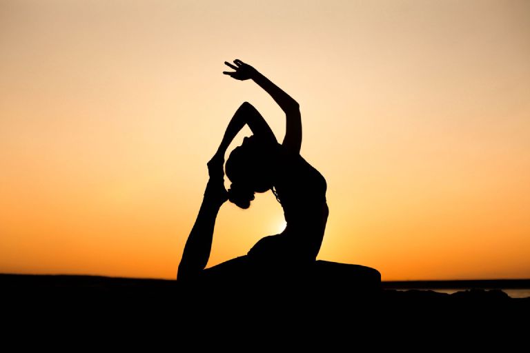 Silhouette of a person in a seated side-stretch yoga pose against an orange sunset sky
